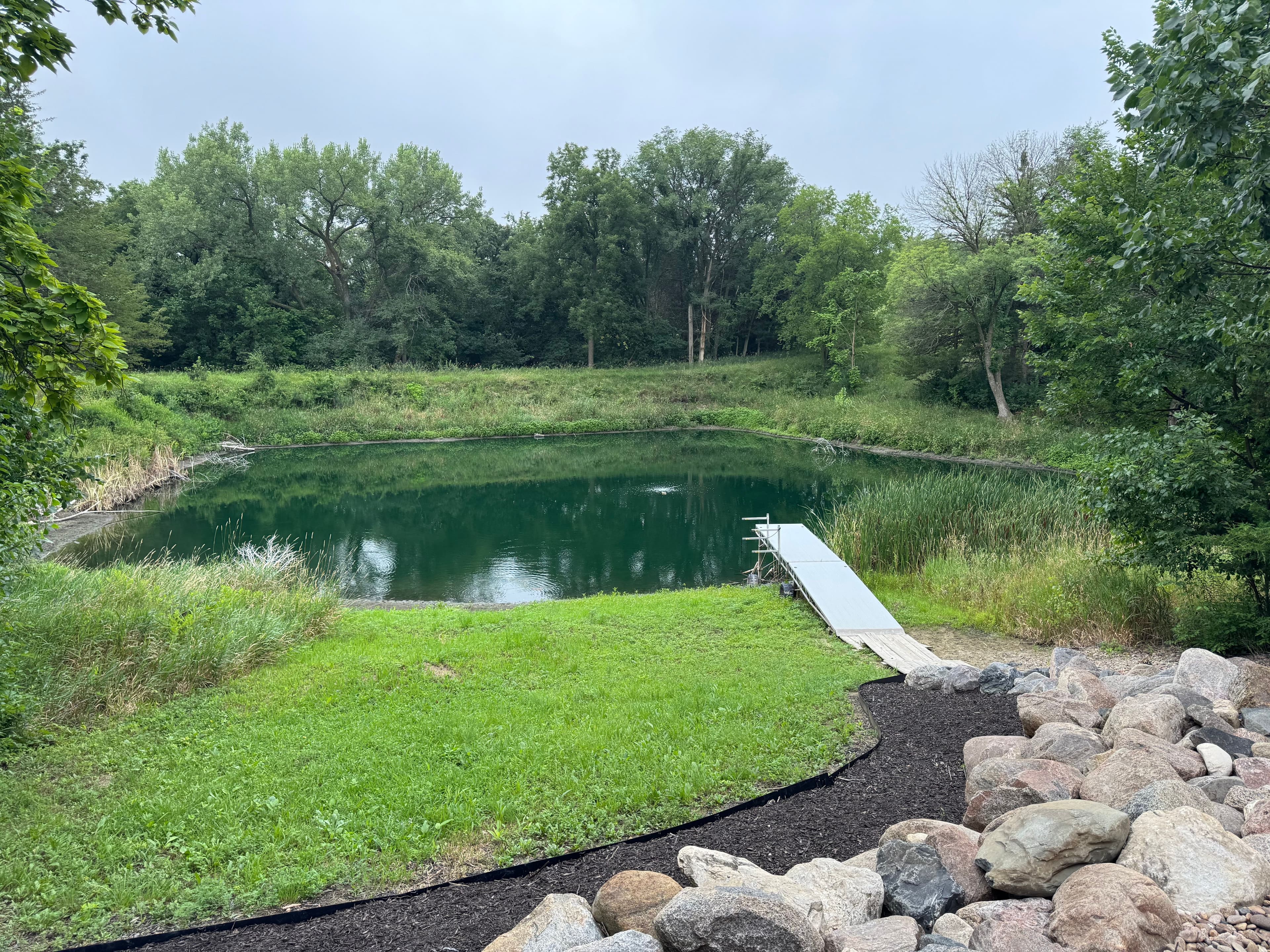 Tranquil pond surrounded by greenery with a small dock and rocky shore.